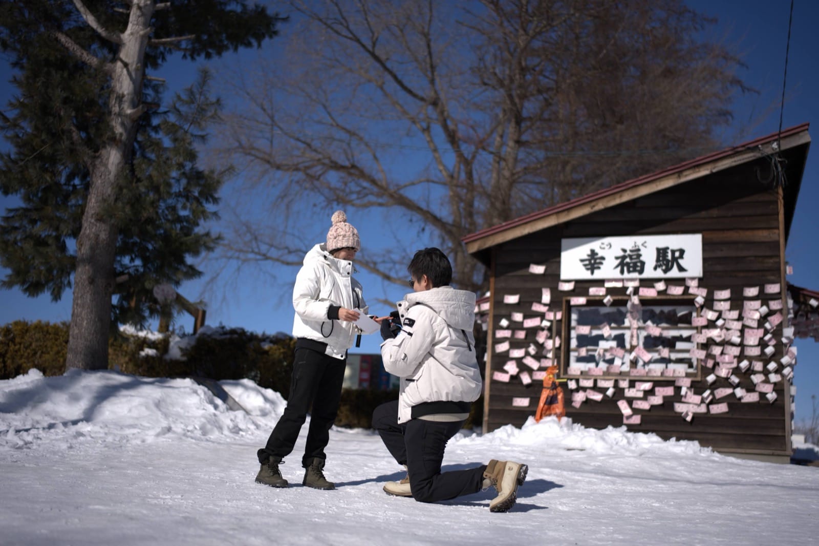Hanny on one knee in the snow proposing to Flora outside a small wooden Hokkaido station hung with wishes.
